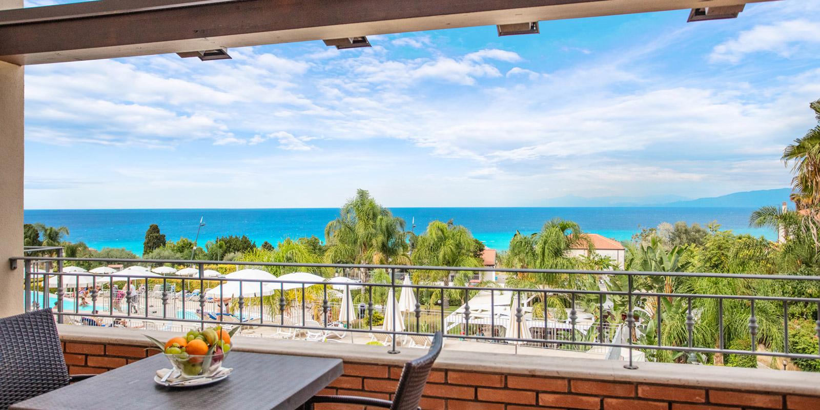 Vista mare da un balcone con tavolo e sedie, circondato da palme e piscina.