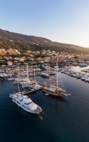 Porto turistico con yacht e barche a vela al tramonto, circondato da colline e case.