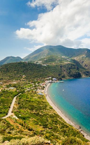 Paesaggio costiero con spiaggia, mare blu e colline verdi sotto un cielo nuvoloso.