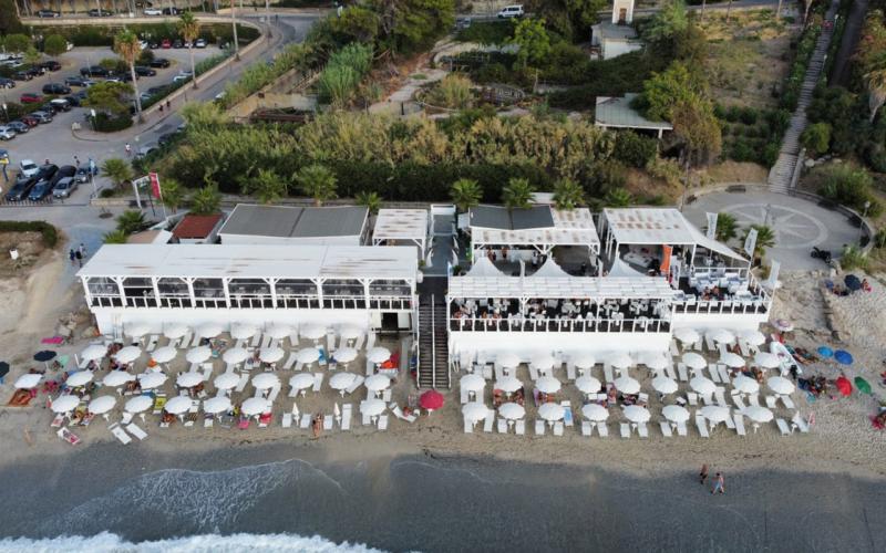 Stabilimento balneare con ombrelloni bianchi su spiaggia e ristorante sopraelevato.